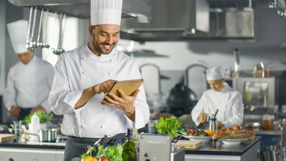 Chef smiling at his ipad while cooks work behind him